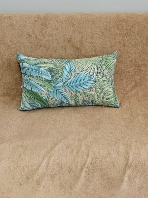 A throw pillow featuring a vibrant tropical leaf print or jungle botanical pattern in shades of teal, turquoise, and various greens. The decorative cushion is placed on a tan or camel-colored sofa with a suede-like texture, set against a light gray wall. This home decor accent provides a bright pop of color and a maximalist or eclectic tropical style for a living space.