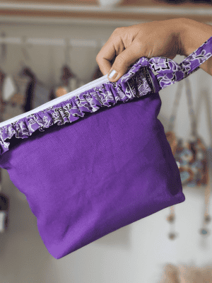 a square zippered pouch or clutch bag with a vibrant solid purple cotton body. The bag is accented with a ruffled trim along the white zipper, made from a black and white batik fabric featuring a floral or vine print. The bag also has a small wrist strap made from the same batik print fabric. This fashion accessory provides a bright pop of color and a bohemian or eclectic style, shown in a retail or closet setting.