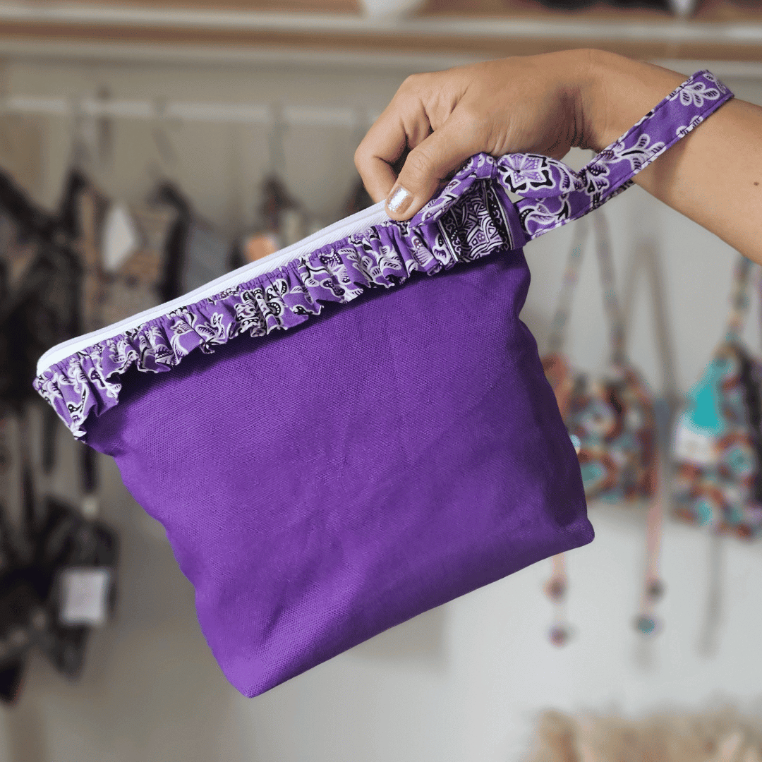 a square zippered pouch or clutch bag with a vibrant solid purple cotton body. The bag is accented with a ruffled trim along the white zipper, made from a black and white batik fabric featuring a floral or vine print. The bag also has a small wrist strap made from the same batik print fabric. This fashion accessory provides a bright pop of color and a bohemian or eclectic style, shown in a retail or closet setting.