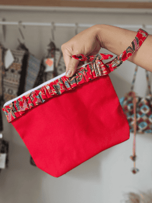 A vibrant zippered pouch bag or clutch with a solid red cotton body. The bag is decorated with a wide, ruffled trim and interior lining made from a red batik fabric with a multicolor floral print in shades of gold, teal, and brown. The bag is shown open and includes a matching batik fabric wrist strap or pull tab. It is resting on a dark, rustic wooden surface, with a logo and dried pampas grass in the background, highlighting an eclectic fashion accessory perfect for travel or a statement handbag.