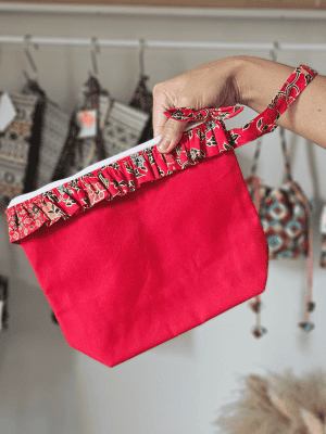 A vibrant zippered pouch bag or clutch with a solid red cotton body. The bag is decorated with a wide, ruffled trim and interior lining made from a red batik fabric with a multicolor floral print in shades of gold, teal, and brown. The bag is shown open and includes a matching batik fabric wrist strap or pull tab. It is resting on a dark, rustic wooden surface, with a logo and dried pampas grass in the background, highlighting an eclectic fashion accessory perfect for travel or a statement handbag.