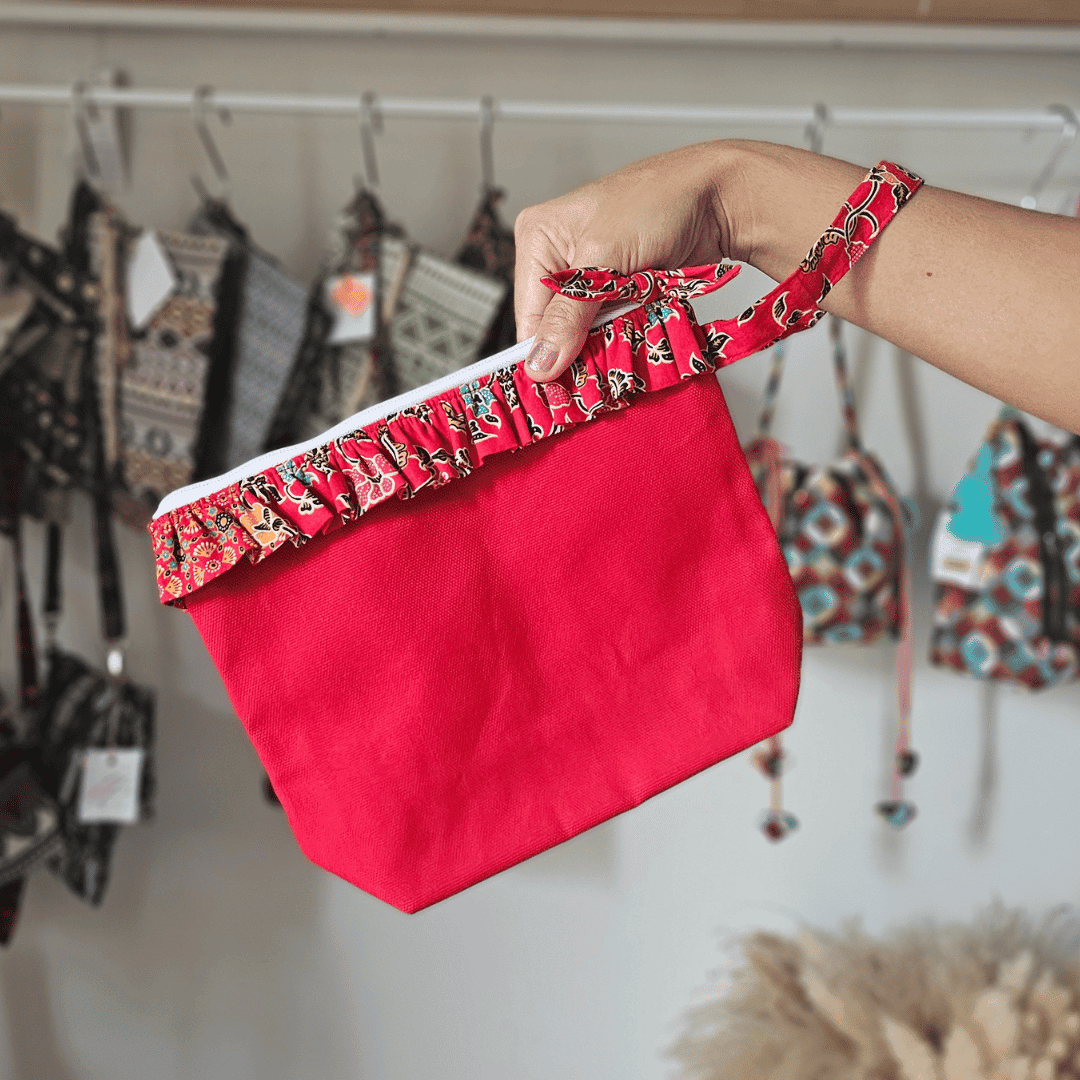 A vibrant zippered pouch bag or clutch with a solid red cotton body. The bag is decorated with a wide, ruffled trim and interior lining made from a red batik fabric with a multicolor floral print in shades of gold, teal, and brown. The bag is shown open and includes a matching batik fabric wrist strap or pull tab. It is resting on a dark, rustic wooden surface, with a logo and dried pampas grass in the background, highlighting an eclectic fashion accessory perfect for travel or a statement handbag.