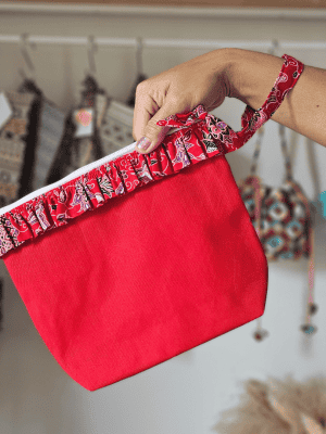 A vibrant zippered pouch bag or clutch with a solid bright red cotton body. The bag is decorated with a wide, ruffled trim and interior lining made from a red batik fabric with a multicolor floral print in shades of purple, white, and dark red. The bag is shown open and includes a matching batik fabric wrist strap. It rests on a dark, rustic wooden surface, with a logo and dried pampas grass in the background, highlighting an eclectic fashion accessory perfect for travel or a statement handbag.