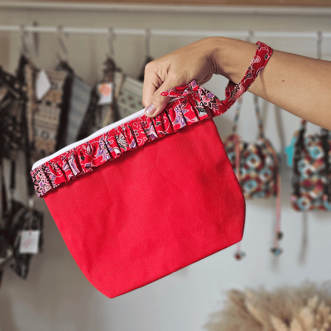 A vibrant zippered pouch bag or clutch with a solid bright red cotton body. The bag is decorated with a wide, ruffled trim and interior lining made from a red batik fabric with a multicolor floral print in shades of purple, white, and dark red. The bag is shown open and includes a matching batik fabric wrist strap. It rests on a dark, rustic wooden surface, with a logo and dried pampas grass in the background, highlighting an eclectic fashion accessory perfect for travel or a statement handbag.