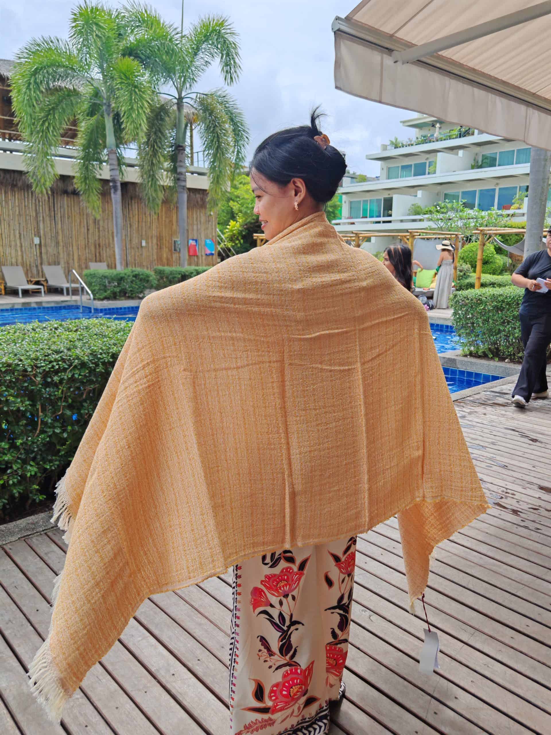 Woman from behind, wearing a floral print outfit and a large light orange woven throw, standing on a wooden deck by a swimming pool.