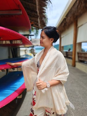 Woman in a cream-colored shawl or throw looking sideways while touching a pink surfboard or paddleboard on a rack near a beach resort.