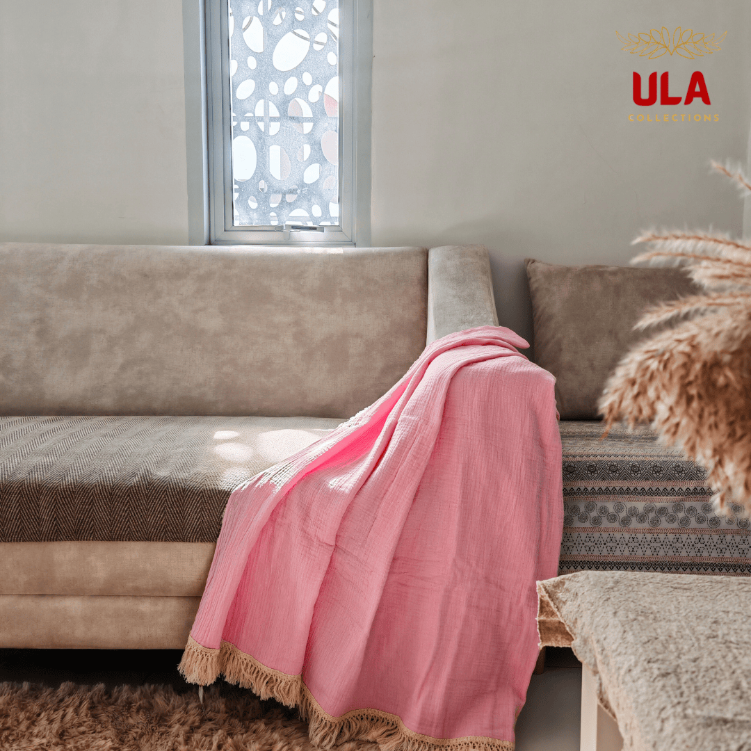 Pink-colored Cotton candy double layer salu throw blanket with tassel trim on a neutral couch, minimalist living room aesthetic by ULA Collections.