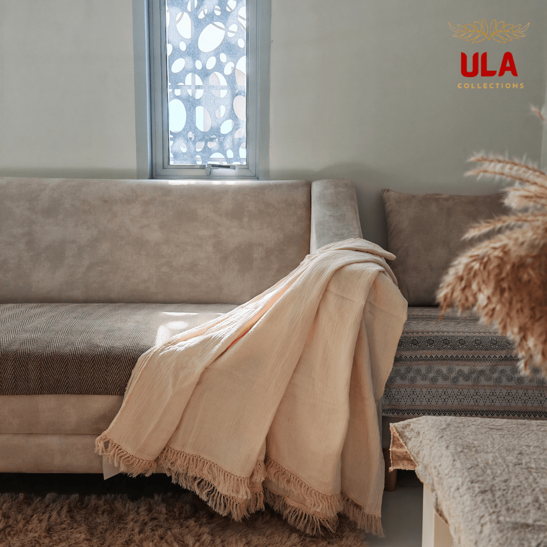 Cream-colored Marshmallow double layer salu throw blanket with tassel trim on a neutral couch, minimalist living room aesthetic by ULA Collections.
