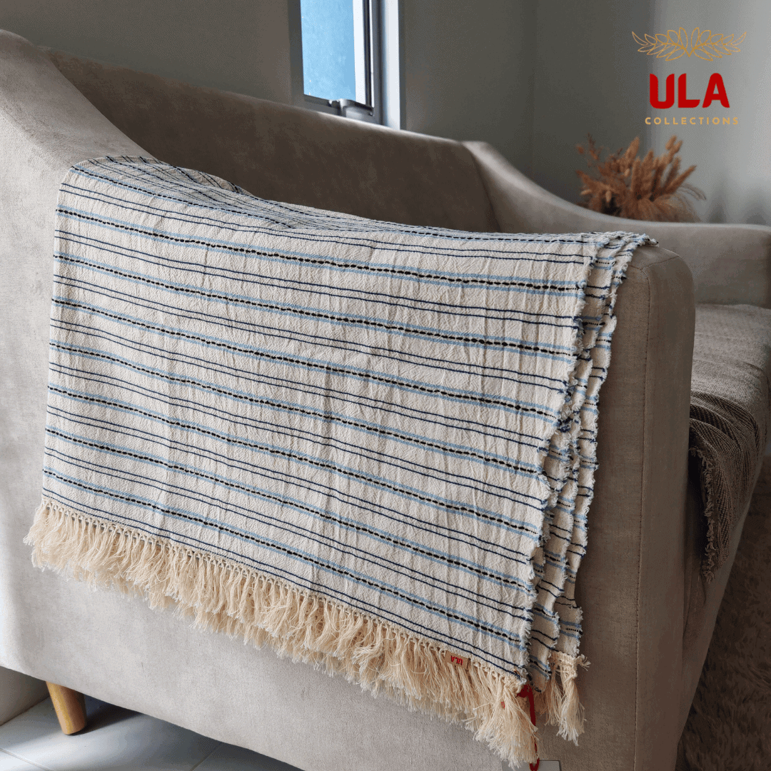 Close-up of ULA Collections light cream throw blanket with navy and light blue stripes and thick fringe, folded over the arm of a curved light tan sofa.
