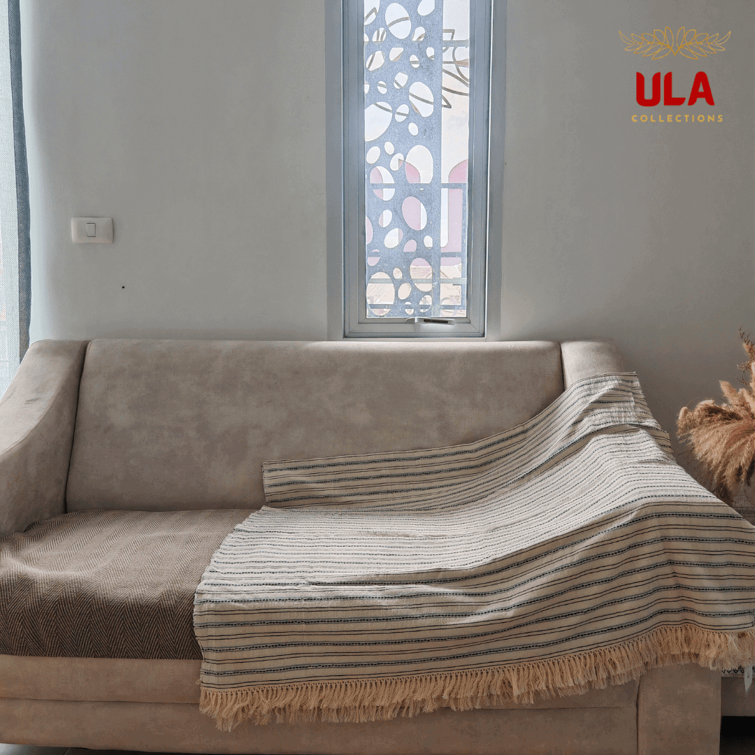 light cream throw blanket with fine navy and light blue horizontal stripes and fringe, draped over a neutral beige sofa.