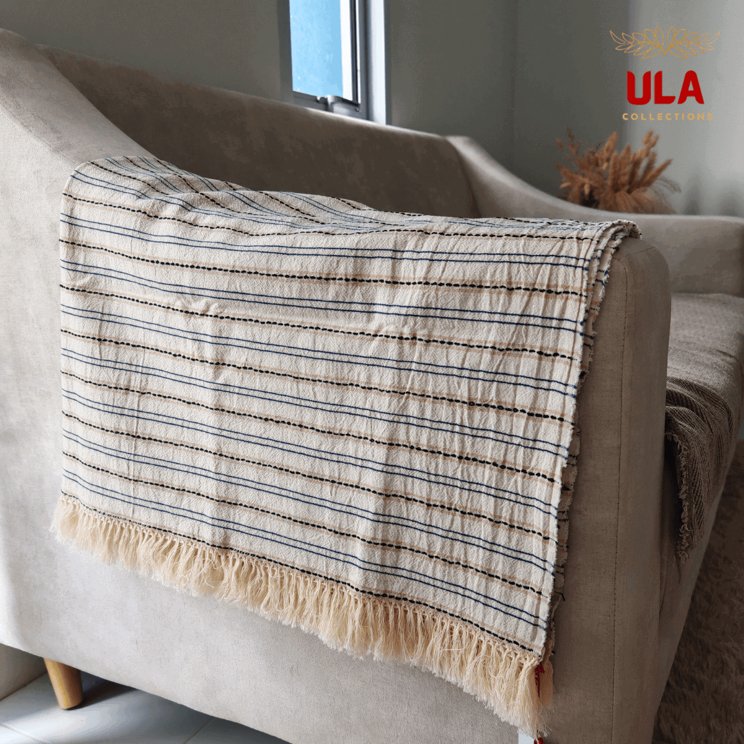 Close-up of ULA Collections light cream throw blanket with black and tan stripes and thick fringe, folded over the arm of a curved light tan sofa.