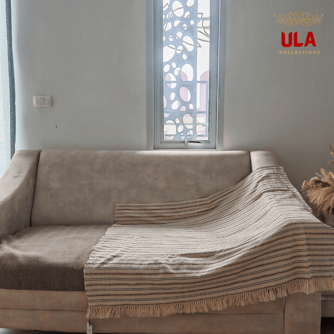 light cream throw blanket with fine black and tan horizontal stripes and fringe, draped over a neutral beige sofa.