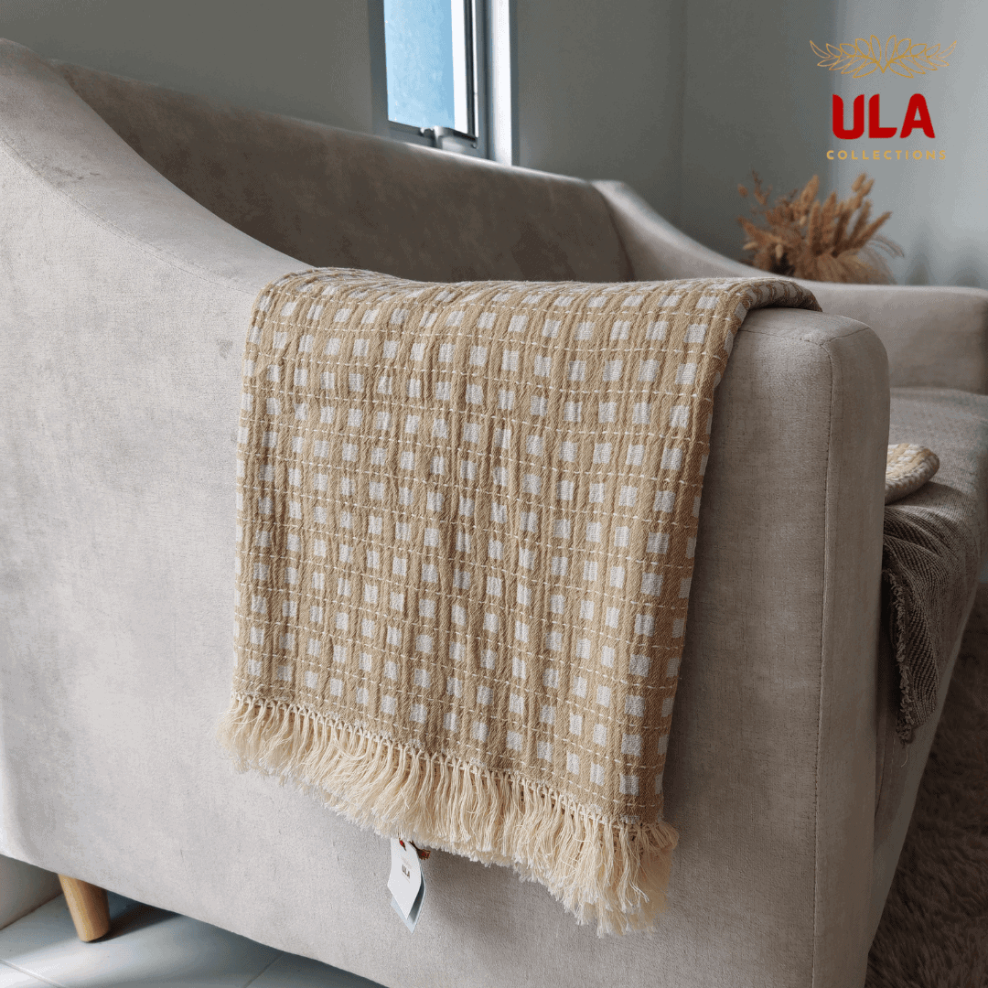 Close-up of ULA Collections light khaki/tan throw blanket with white checkered weave and cream fringe, folded over the arm of a curved beige sofa.