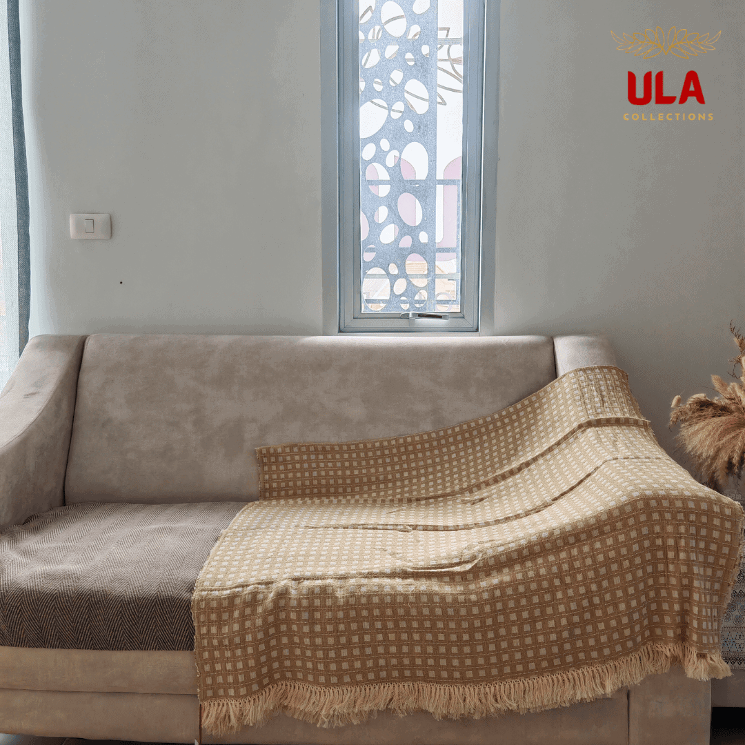 light khaki woven throw blanket with white square or windowpane check pattern and fringe, draped over a neutral beige sofa.