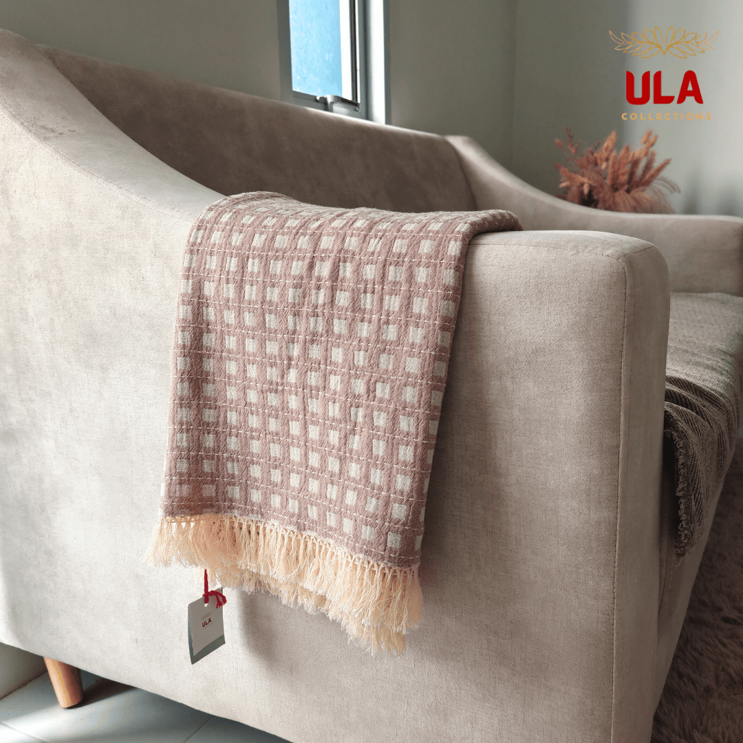 Close-up of ULA Collections plum taupe/pinkish brown throw blanket with white checkered weave and cream fringe, folded over the arm of a curved beige sofa.