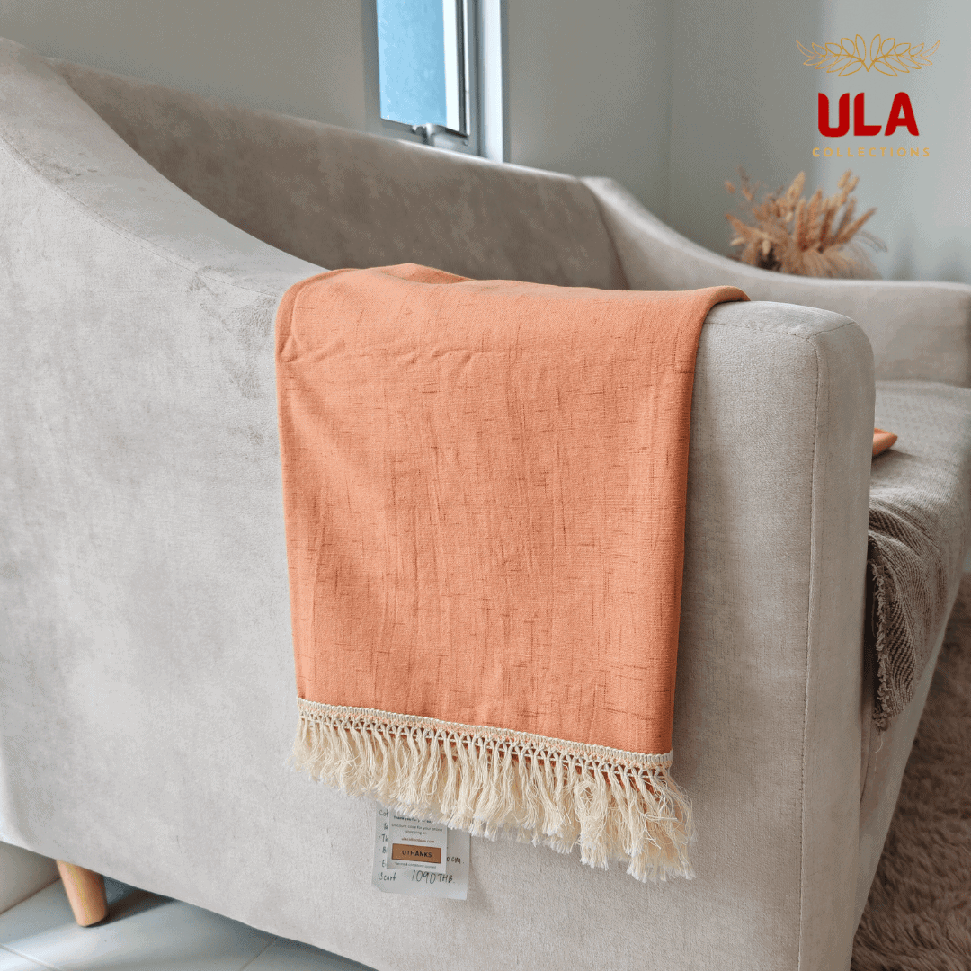 Close-up of ULA Collections burnt orange textured throw blanket with white fringe, draped over the arm of a beige sofa, showing product tag.