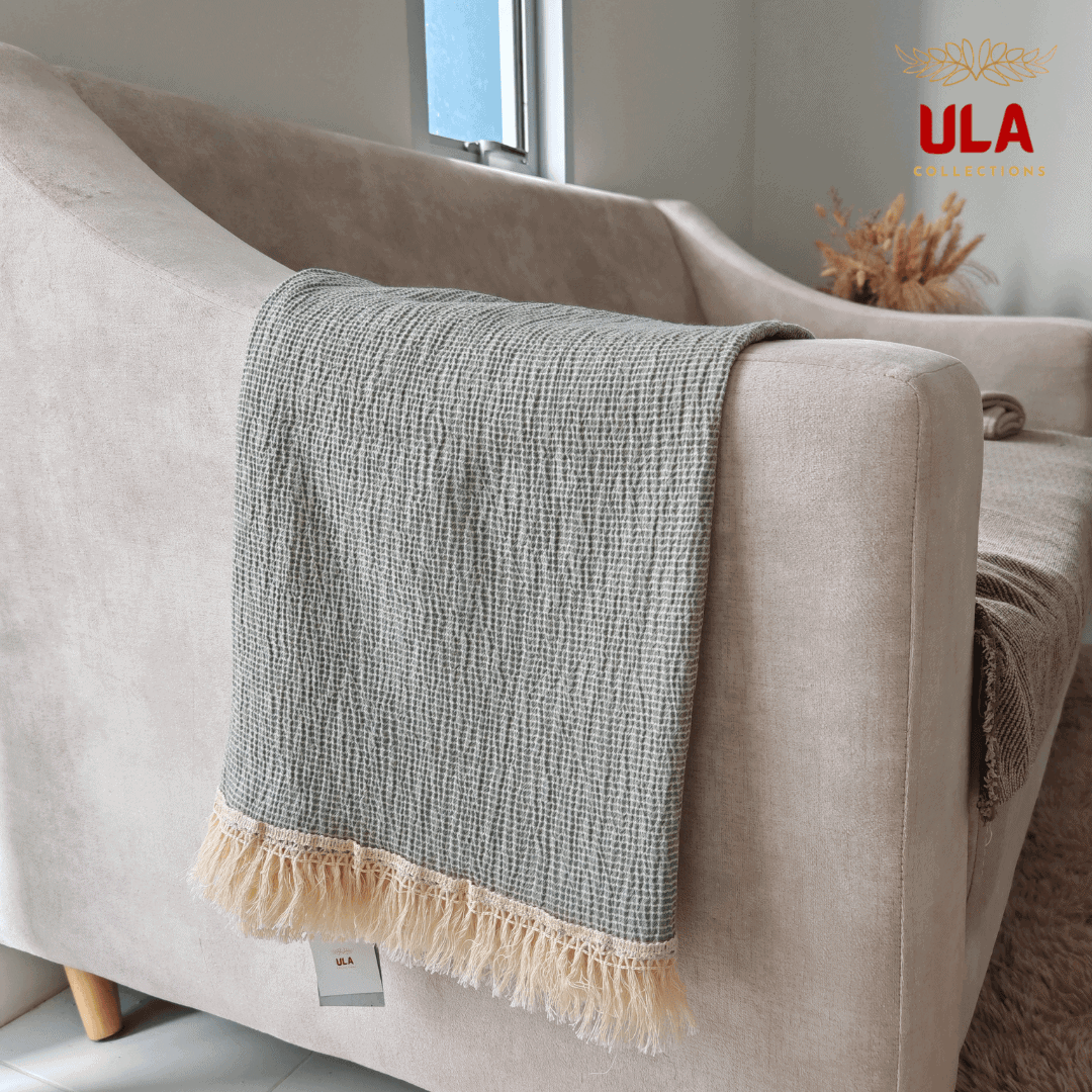 Close-up of ULA Collections solid sage green throw blanket showing crinkled texture and light fringe, folded over the arm of a light tan sofa.
