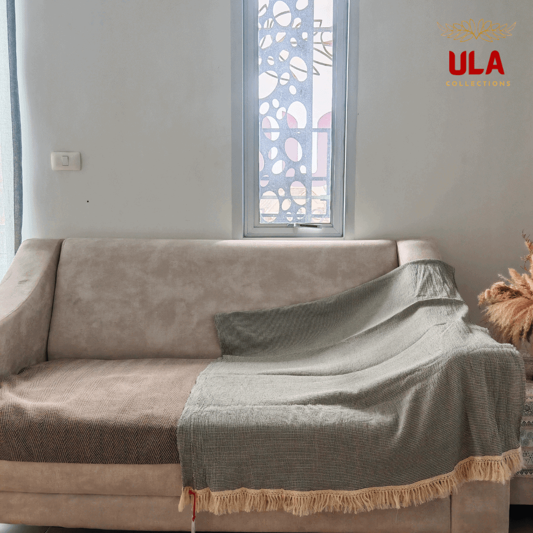 solid sage green textured throw blanket with light fringe, draped over a neutral beige sofa with a brown herringbone cushion cover.