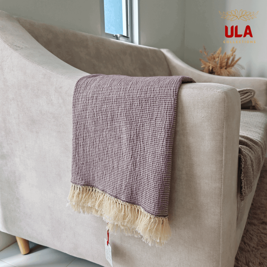 Close-up of ULA Collections solid plum throw blanket showing crinkled texture and light fringe, folded over the arm of a light tan sofa.
