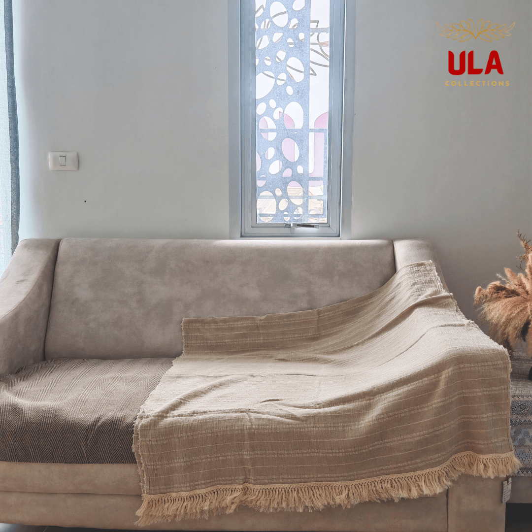 natural tan throw blanket with stripe weave and fringe, made from a silk, hemp, linen, and cotton blend, draped over a beige sofa with a visible section of brown herringbone fabric.