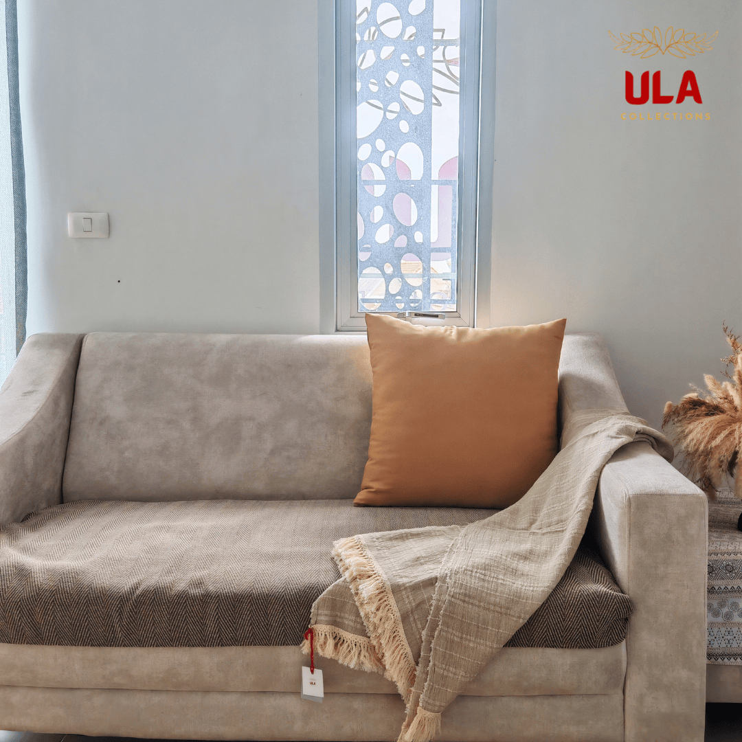 natural tan throw blanket, made from a silk, hemp, linen, and cotton blend, draped over the arm of a beige sofa with an orange accent pillow.