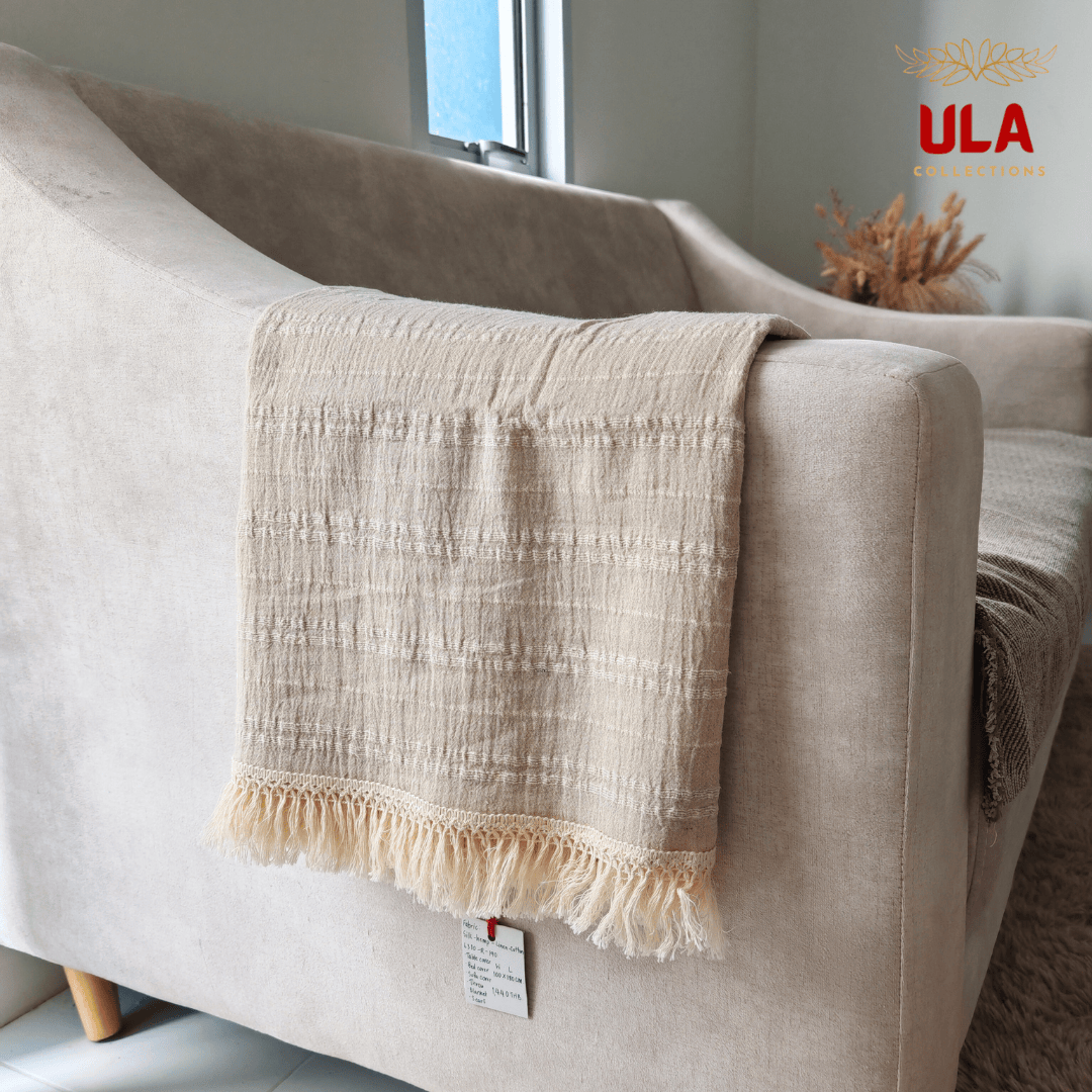 Close-up of a natural beige, striped throw blanket with fringe, made from a silk, hemp, linen, and cotton blend, draped over the arm of a light tan sofa.
