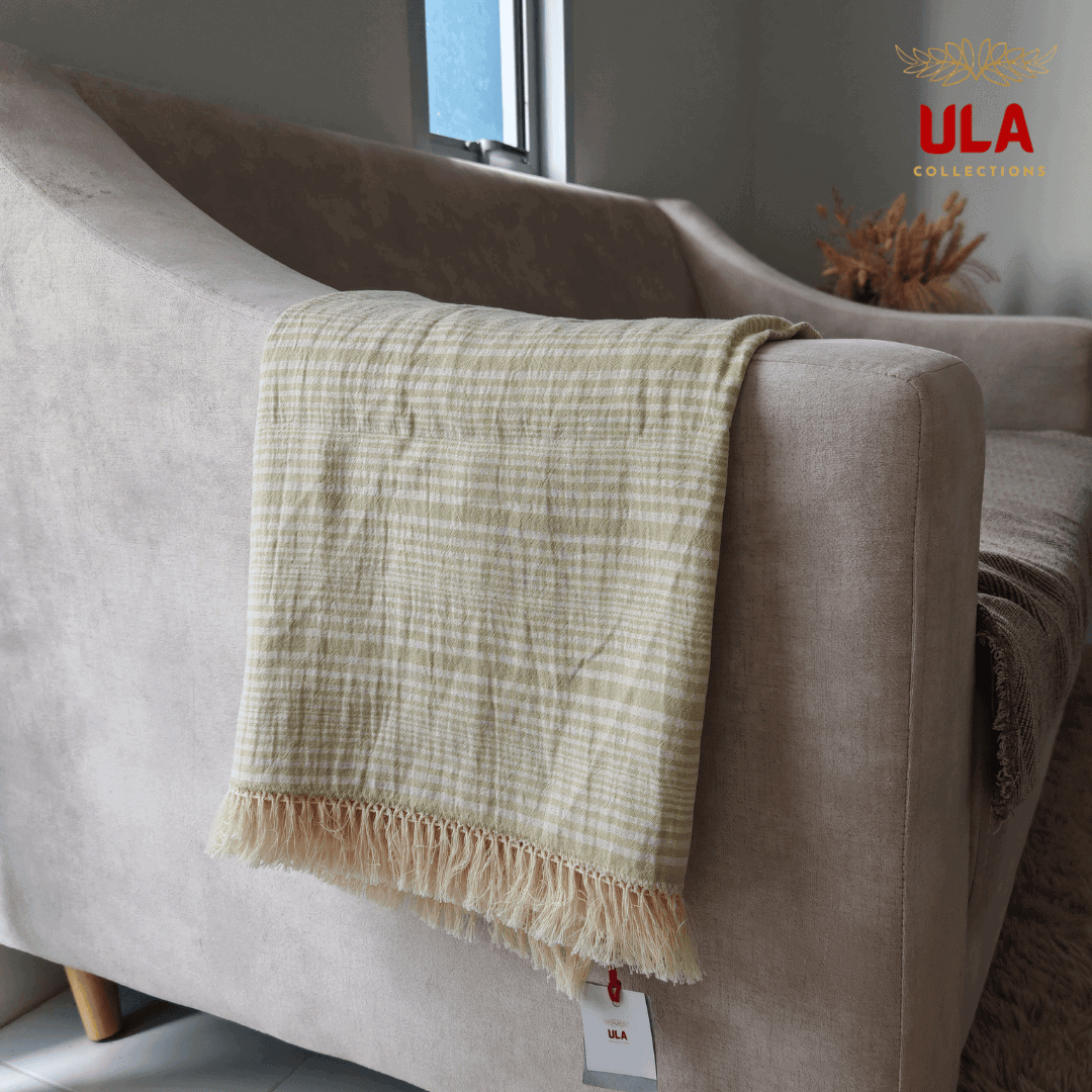 Close-up of ULA Collections light cream and pale green striped throw blanket with fringe, folded over the arm of a curved light tan sofa.
