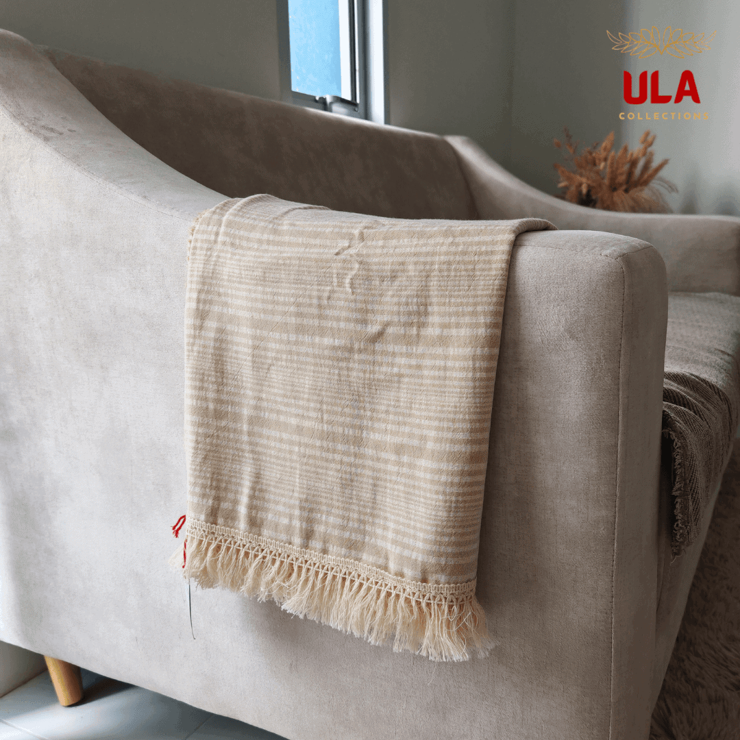 Close-up of ULA Collections light khaki and cream striped throw blanket with fringe, folded over the arm of a curved light tan sofa.