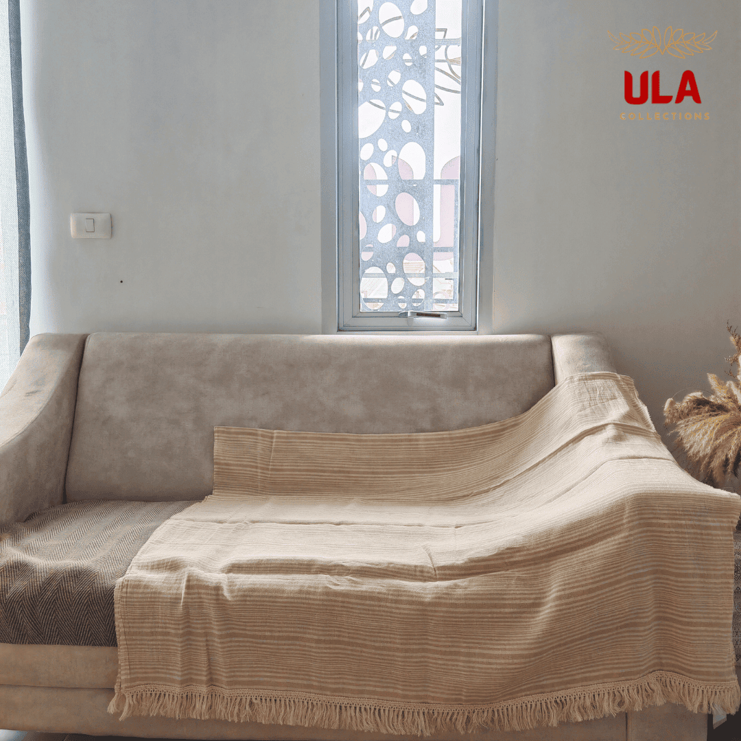 light khaki woven throw blanket with fine cream horizontal stripes and thick fringe, draped over a neutral beige sofa.