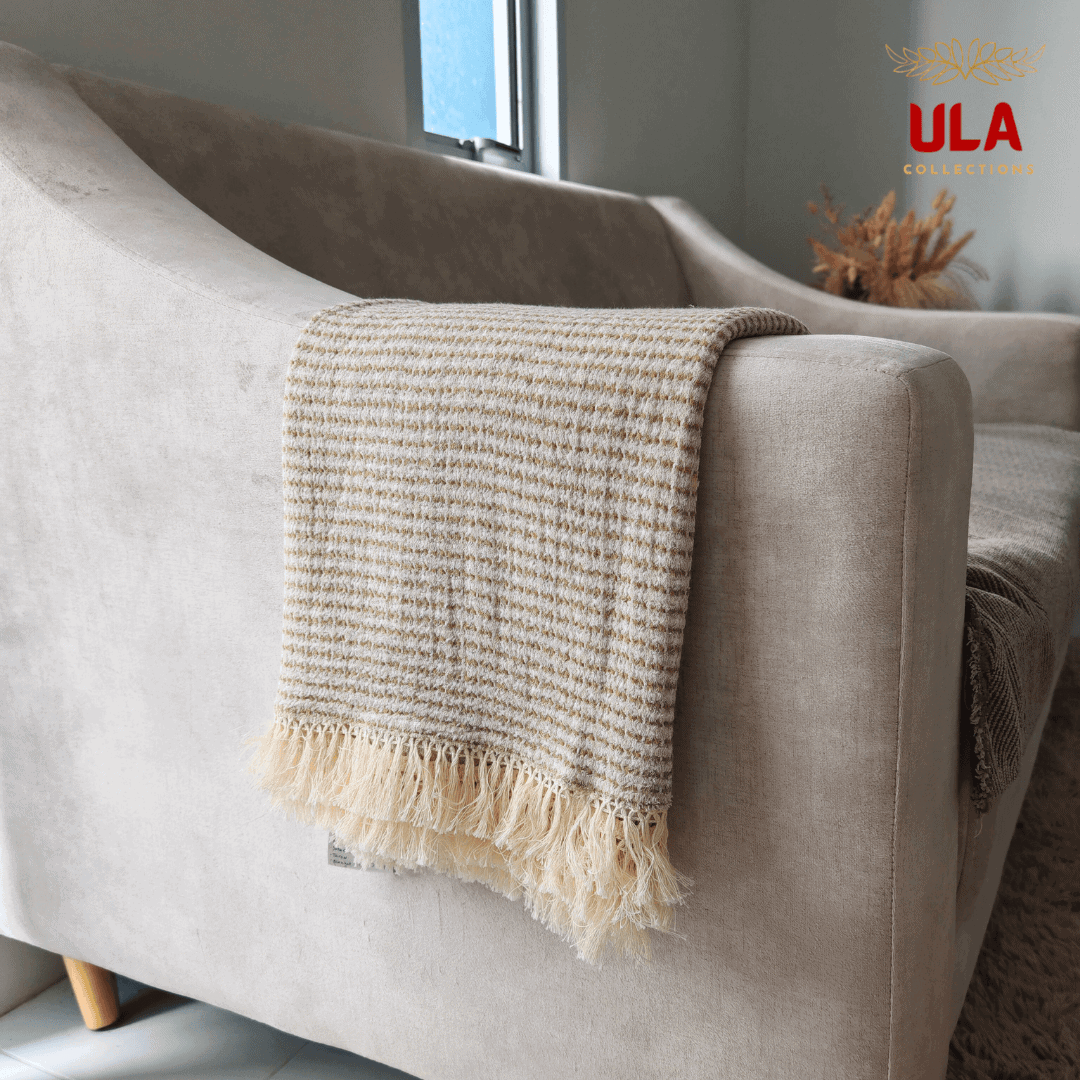 Close-up of ULA Collections cream and olive green striped throw blanket with fringe, folded over the curved arm of a light tan sofa, showing texture.