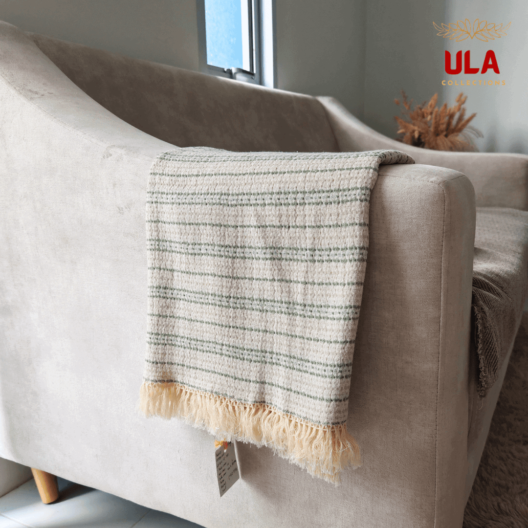 Close-up of ULA Collections cream and sage green striped throw blanket with fringe, folded over the rounded arm of a light tan sofa, showing the weave.