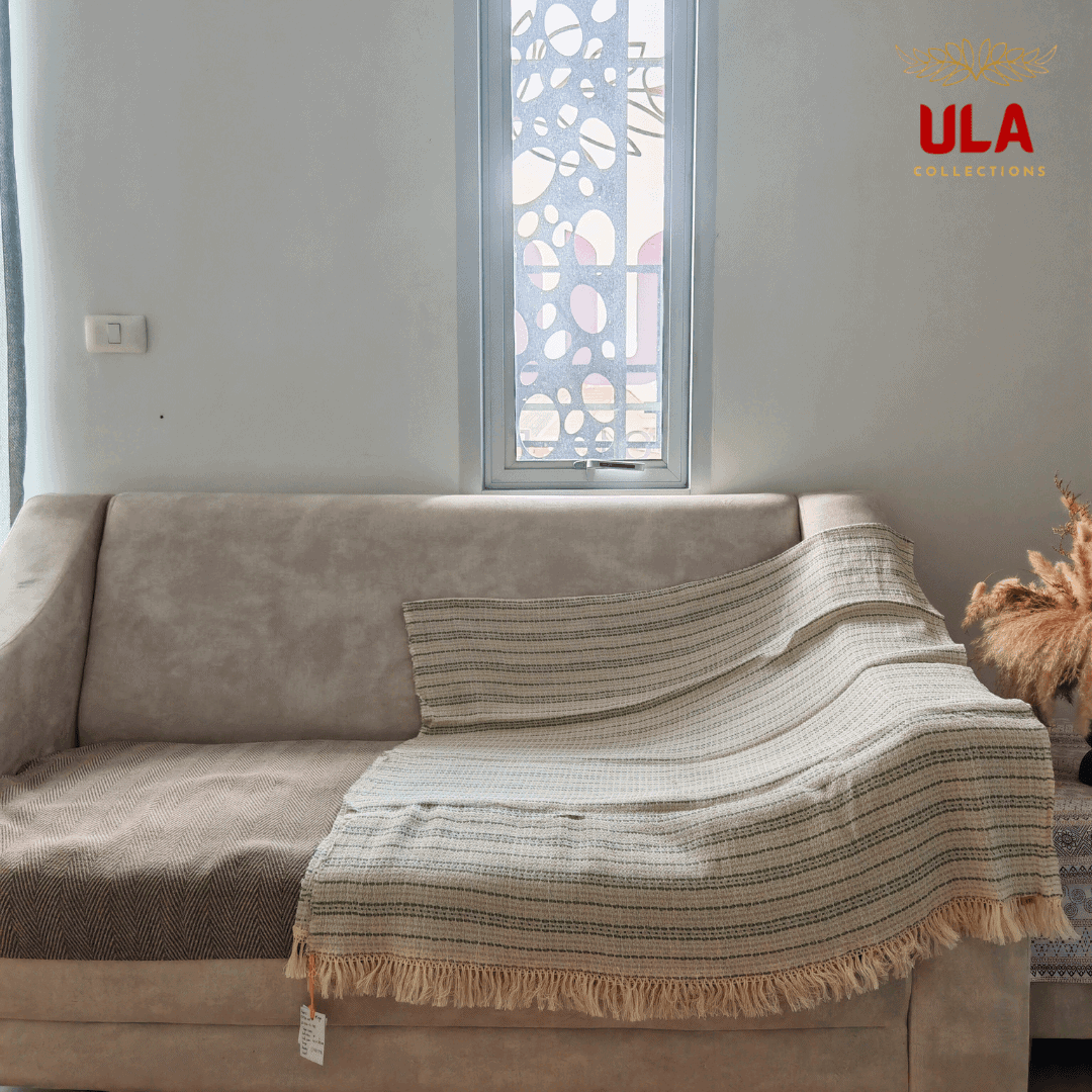 striped woven throw blanket in cream with thin sage green horizontal stripes and fringe, draped over a neutral beige sofa.