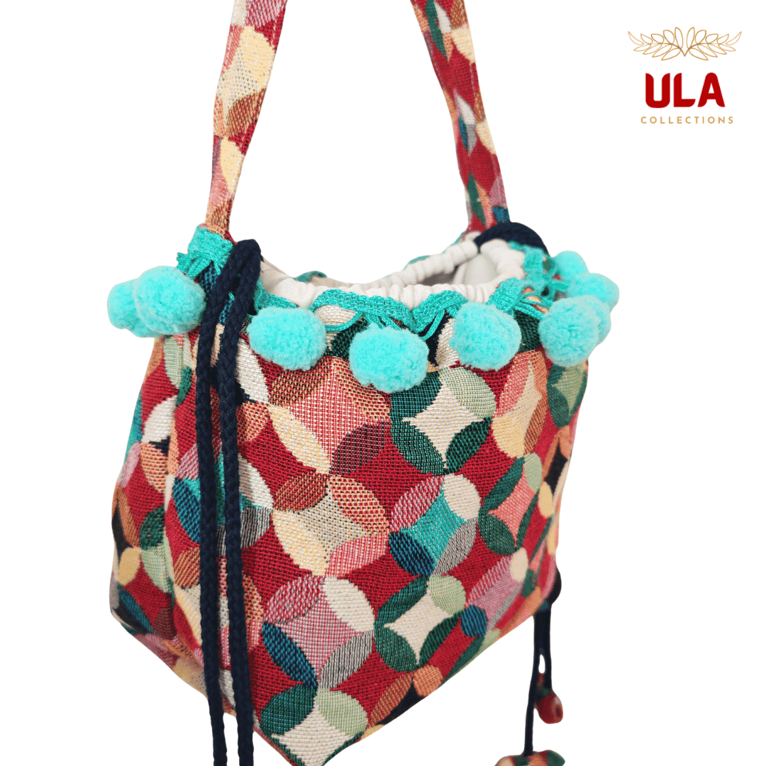 handmade woven drawstring bucket bag with a colorful patchwork/circle pattern in red, green, and cream, featuring teal pom-poms.
