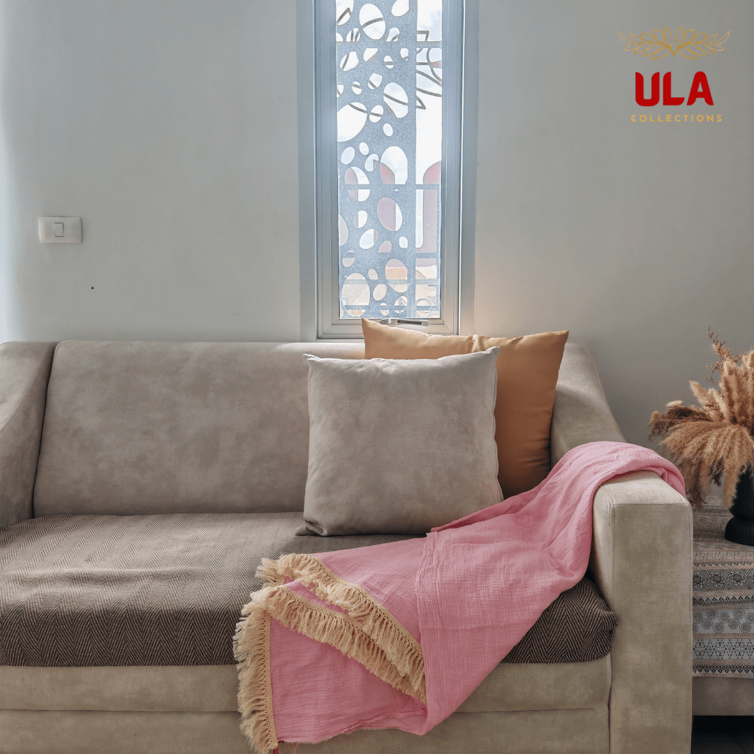 Pink crinkle throw blanket with fringe, casually draped over a beige sofa next to gray and orange accent pillows.