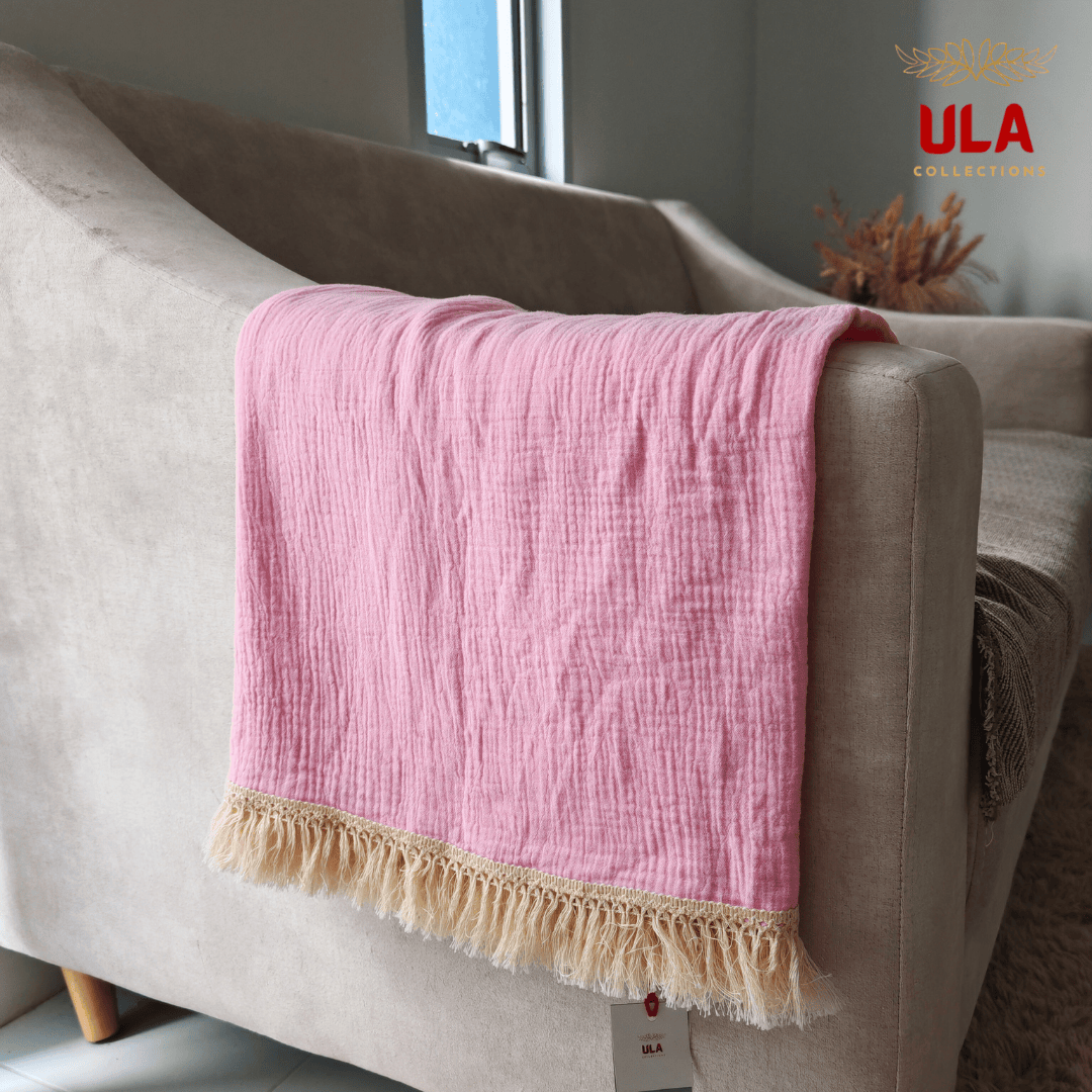 Close-up of ULA Collections bright pink throw blanket showing crinkled/gauze texture and thick cream fringe, folded over the arm of a light tan sofa.