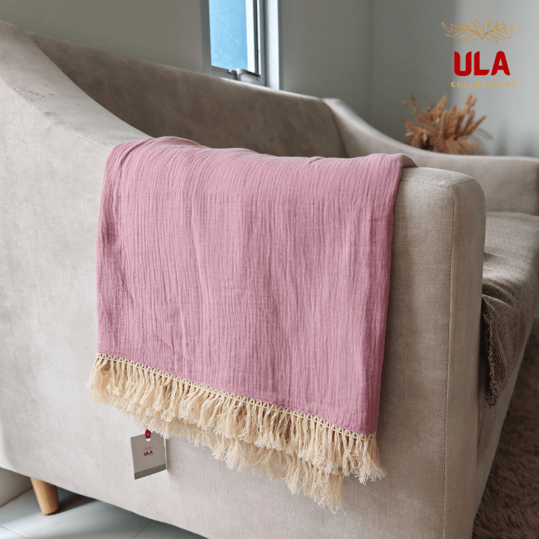 Close-up of ULA Collections rose pink throw blanket showing crinkled/gauze texture and thick cream fringe, folded over the arm of a light tan sofa.