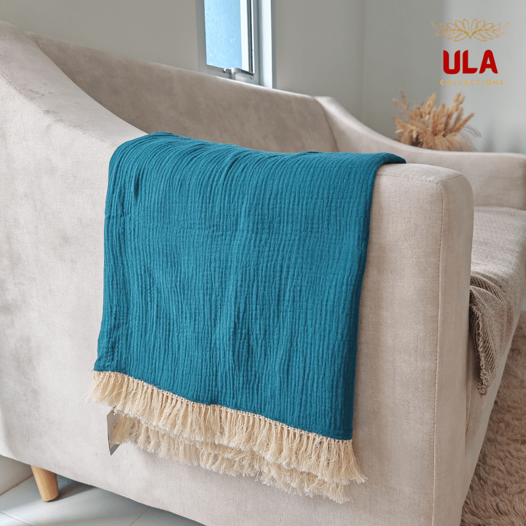 Close-up of ULA Collections vibrant teal throw blanket showing crinkled/gauze texture and thick cream fringe, folded over the arm of a light tan sofa.