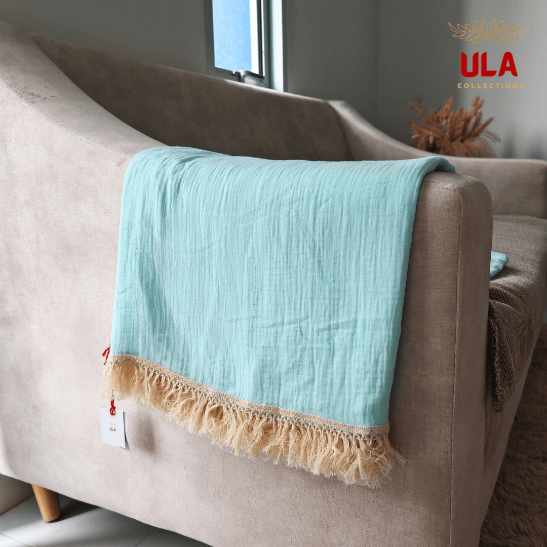 Close-up of ULA Collections light blue throw blanket showing crinkled/gauze texture and thick cream fringe, folded over the arm of a light tan sofa.