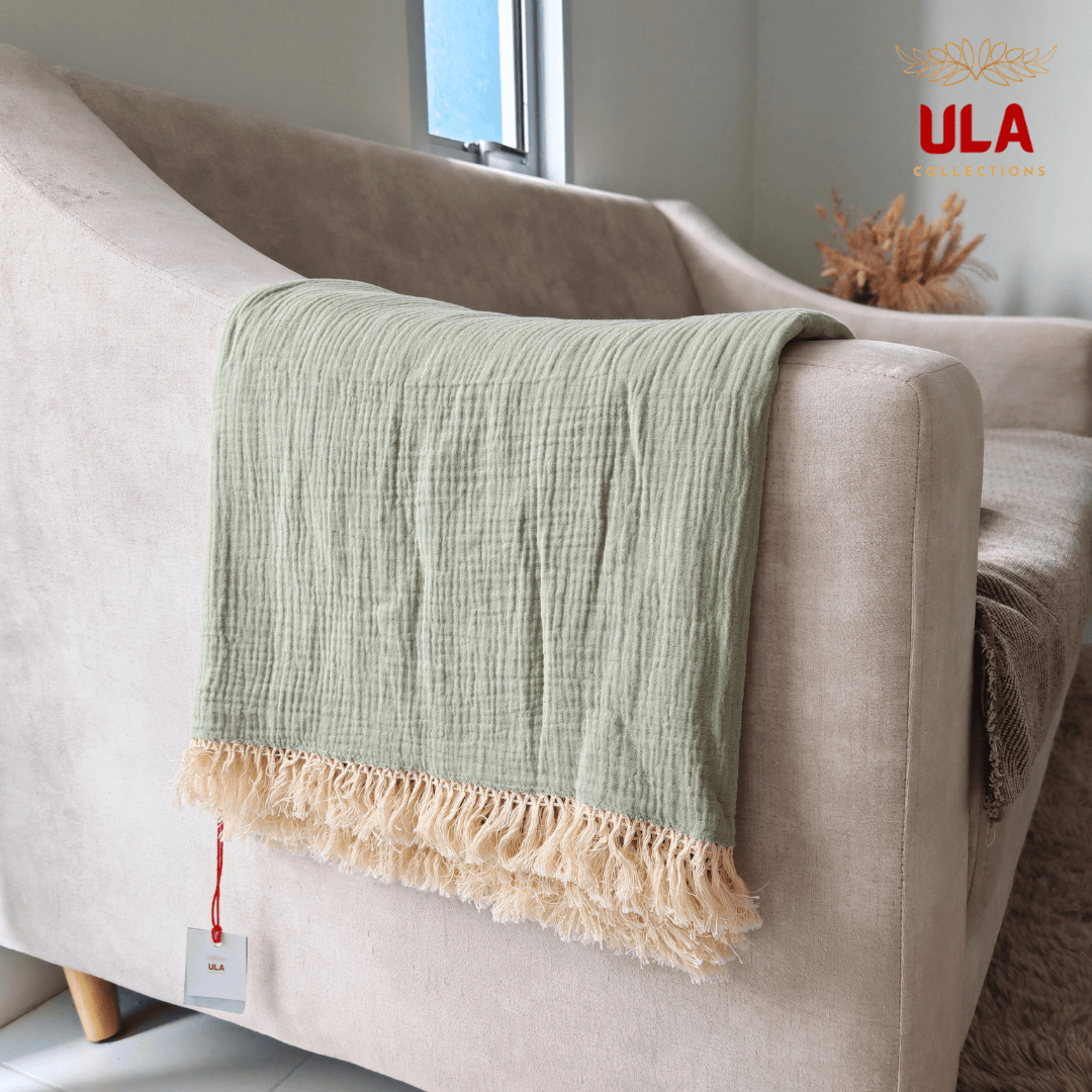 Close-up of ULA Collections pale olive green throw blanket showing crinkled/gauze texture and thick cream fringe, folded over the arm of a light tan sofa.