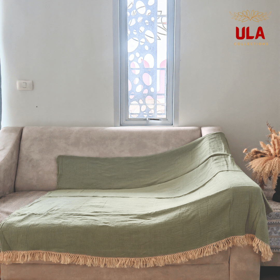 Solid sage green textured throw blanket with thick cream fringe, completely covering a neutral beige sofa.