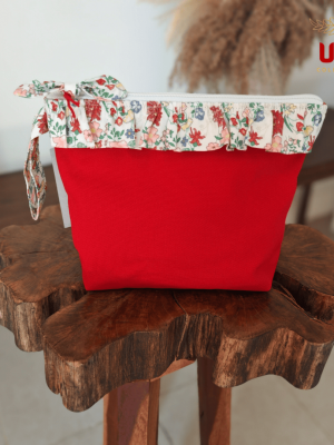 Front view of a vibrant red canvas cosmetic pouch featuring a ruffled floral trim and a matching bow zipper pull, displayed on a rustic wooden stool.