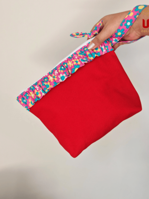 A close-up photograph of a vibrant red canvas pouch being held by a hand. The top of the bag features a colorful, floral ruffle trim in shades of pink, yellow, and turquoise. A matching patterned wristlet strap is looped around the person's wrist. The pouch has a white zipper closure at the top, and the "ULA COLLECTIONS" logo is visible in the upper right corner.