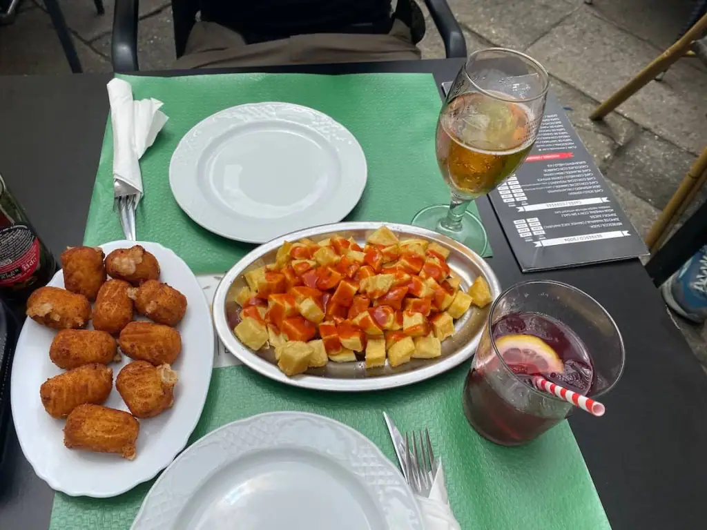 Plate of croquetas, plate of patatas bravas, a glass of tinto de verano, and a glass of beer on a green outdoor table in Santiago de Compostela, Spain.