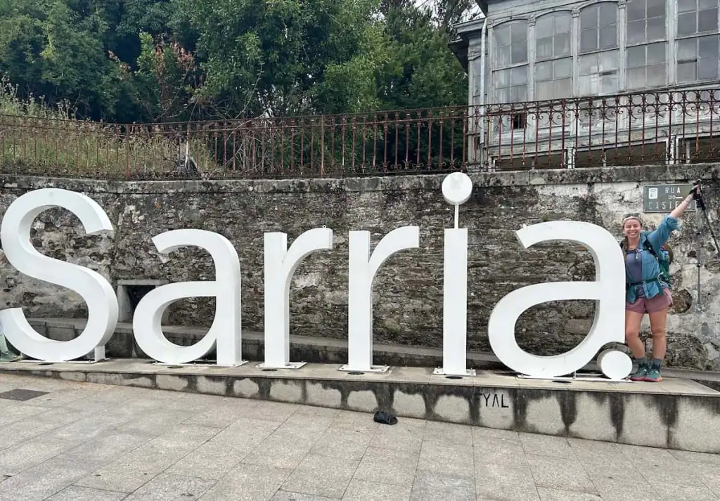 Sign that says "Sarria"and Mikala standing to the right of it smiling and holding her hiking poles in the air in Sarria, Spain on the Camino Francés.