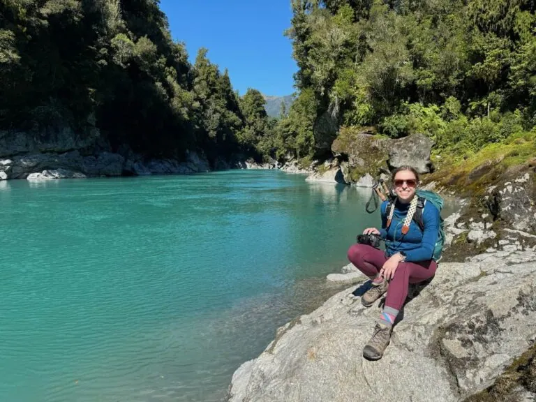 Mikala sitting smiling on a rock in front of Hokitika Gorge in New Zealand