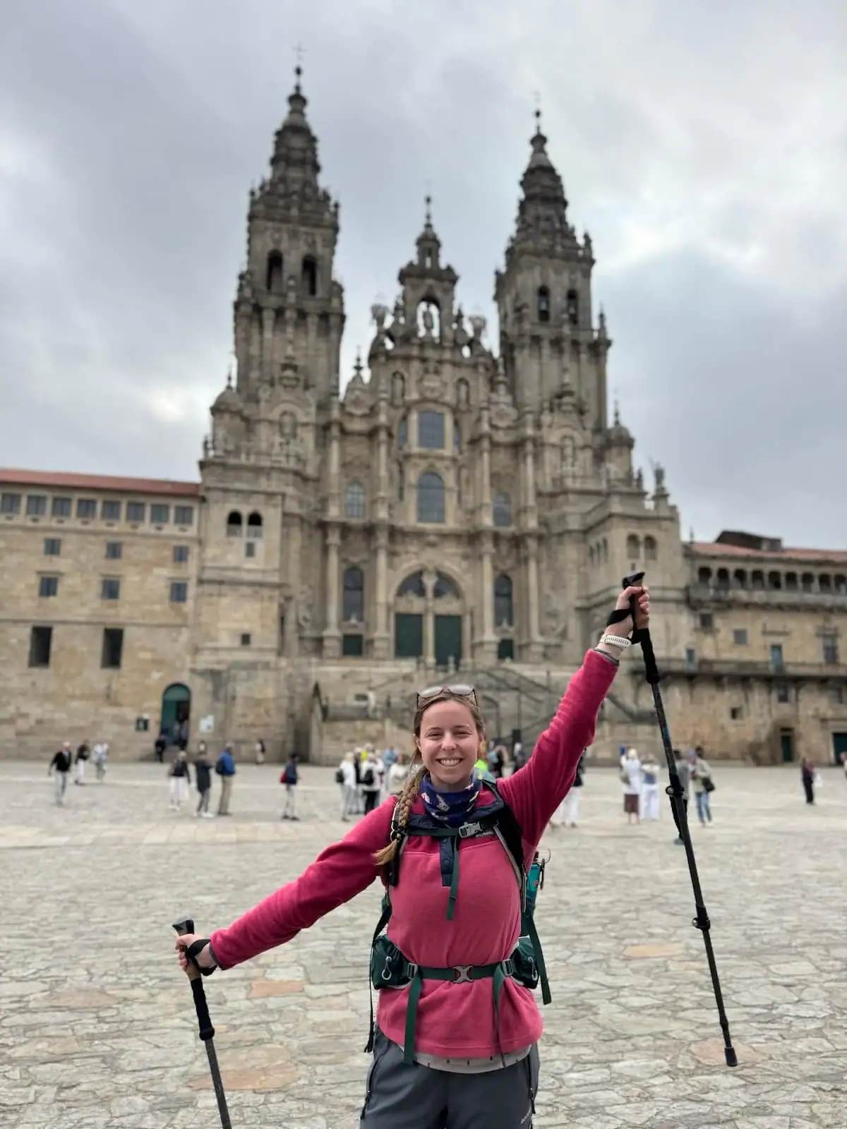 Mikala smiling with her arms up in the air holding her trekking poles in front of the cathedral in Santiago de Compostela after finishing her Portuguese Coastal Camino route stages.