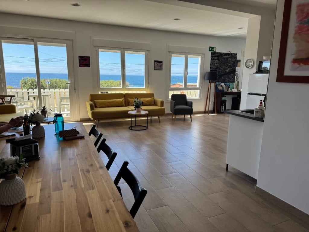 Table with chairs and couches with windows overlooking the ocean at Albergue da Estrela in Viladesuso along the Camino Portugués Coastal route.