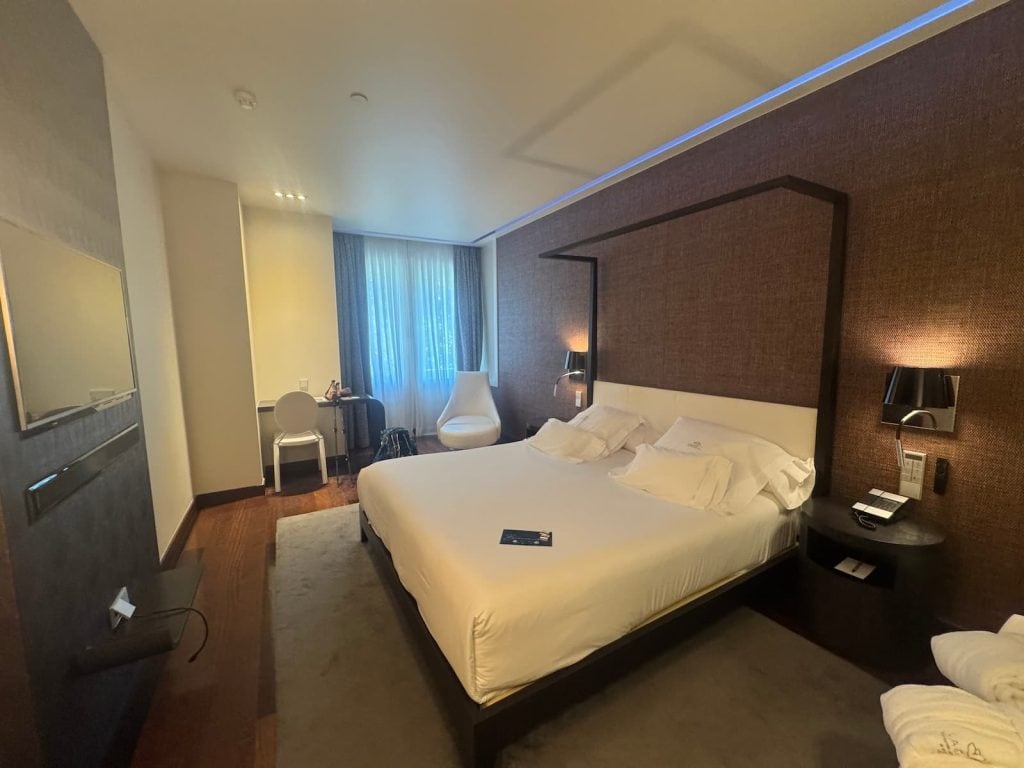 Large white bed in a hotel room with a desk and open window.