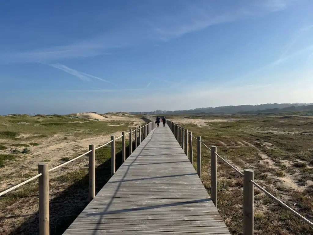 Boardwalk on the Camino Portugués Coastal route stretching ahead with two pilgrims walking along it in the distance, sand dunes on both sides, and sunny clear blue skies above.