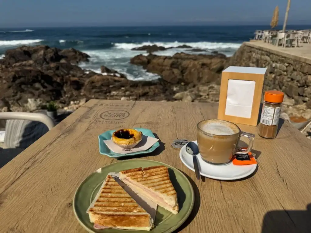 A coffee, toasted ham and cheese sandwich, and pastel de nata on a table with a view of the ocean along the Camino Portugués Coastal route.